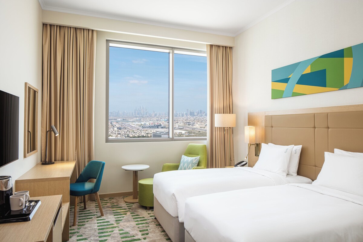 A hotel room with two beds, a desk with a coffee maker, a TV, a view of the city, and a painting on the wall.