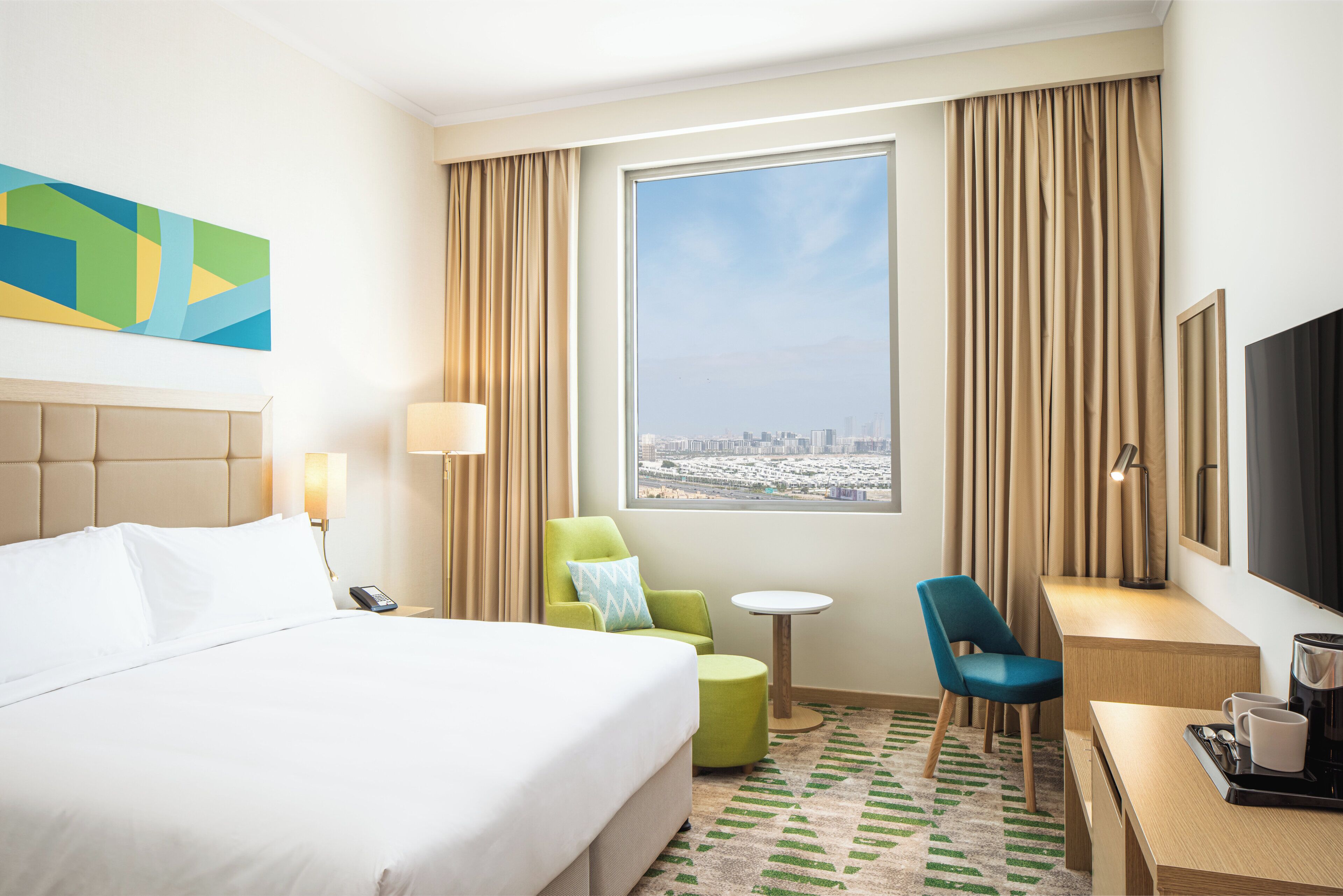 A hotel room with a bed, a desk, a chair, a small table, a TV, and a large window with a city view.