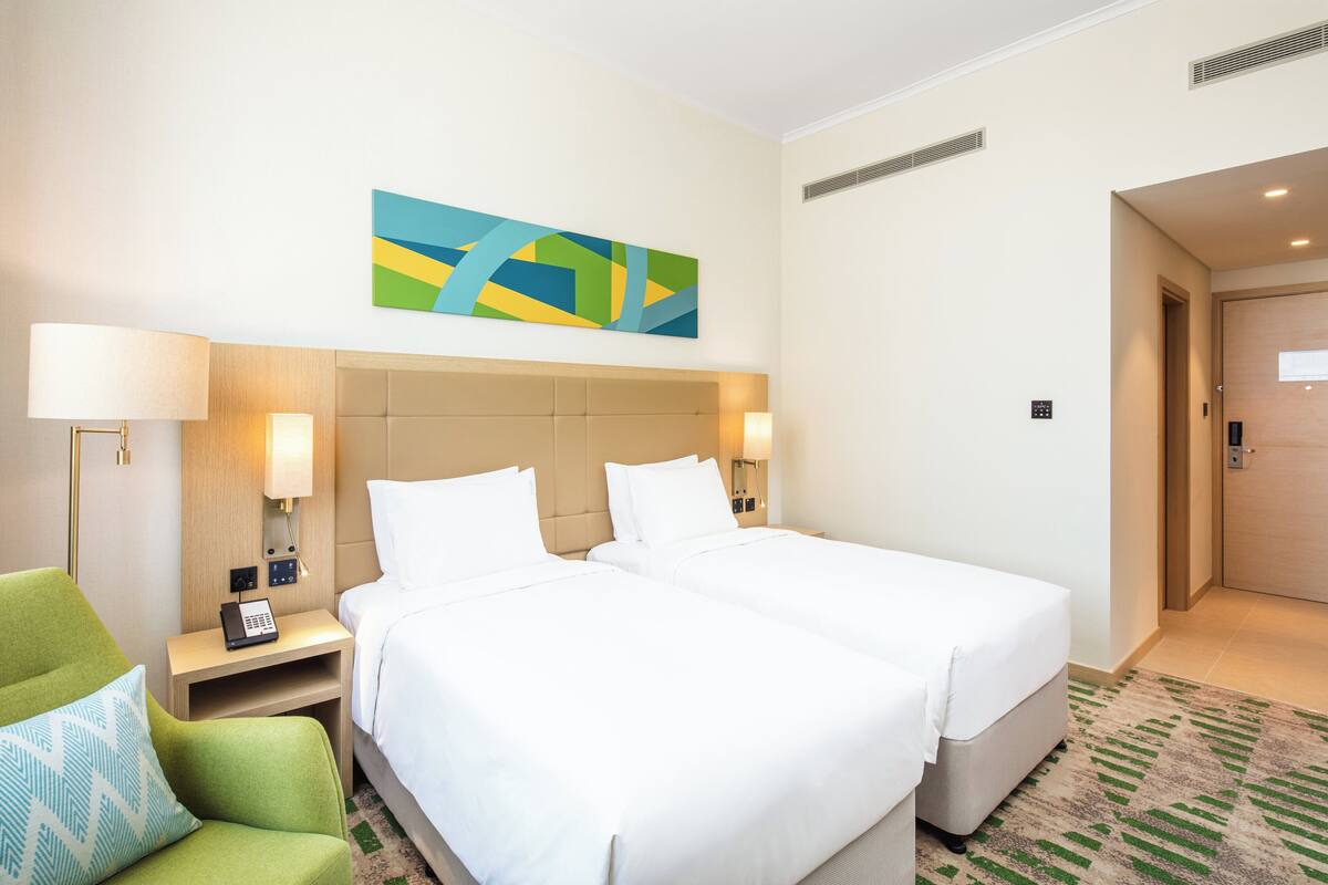 Een hotelkamer met twee bedden, een groene fauteuil, een nachtkastje met een lamp en een schilderij aan de muur.