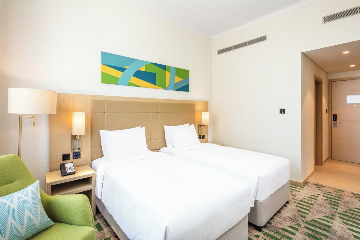 A hotel room with two beds, a green armchair, a bedside table with a lamp, and a painting on the wall.