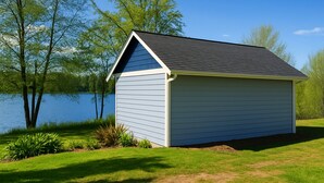 A small, single-story shed with a sloped roof, located beside a body of water with trees in the background.
