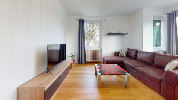 Ein Wohnzimmer mit einem Fernsehschrank aus Holz, einem braunen Ledersofa, einem Couchtisch in Rot und Schwarz, einem Heizkörper und einem Fenster mit Vorhängen.
