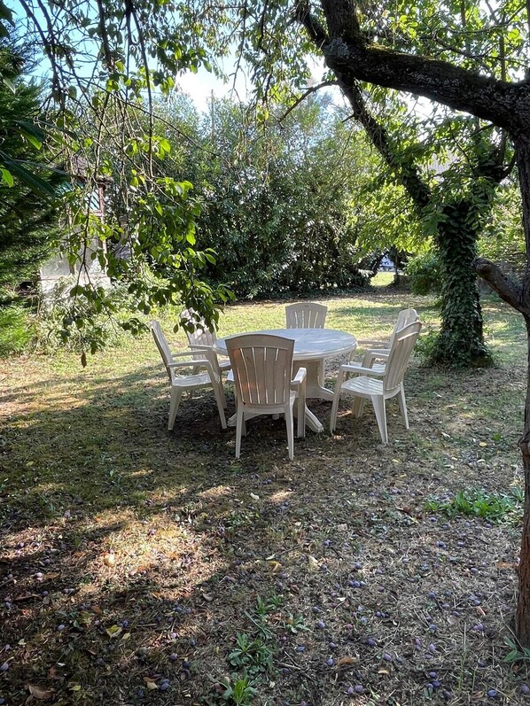 An outdoor dining area with a round table and six chairs, surrounded by trees and grass.