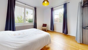Ein Schlafzimmer mit einem Bett, einem Sessel, einem Heizkörper und großen Fenstern mit Vorhängen.