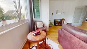 Ein Wohnzimmer mit einer Couch, einem kleinen runden Tisch mit einer Schachtel, einer Lampe und Blick durch ein Fenster.