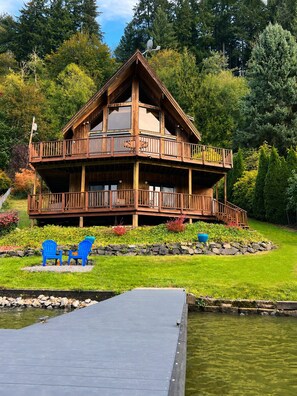 Eine zweistöckige Holzhütte mit Balkon, umgeben von einem angelegten Garten und einem Steg, der ins Wasser führt.