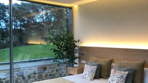 Een moderne slaapkamer met een groot raam, een bed met een beige deken en een nachtkastje met een lamp.