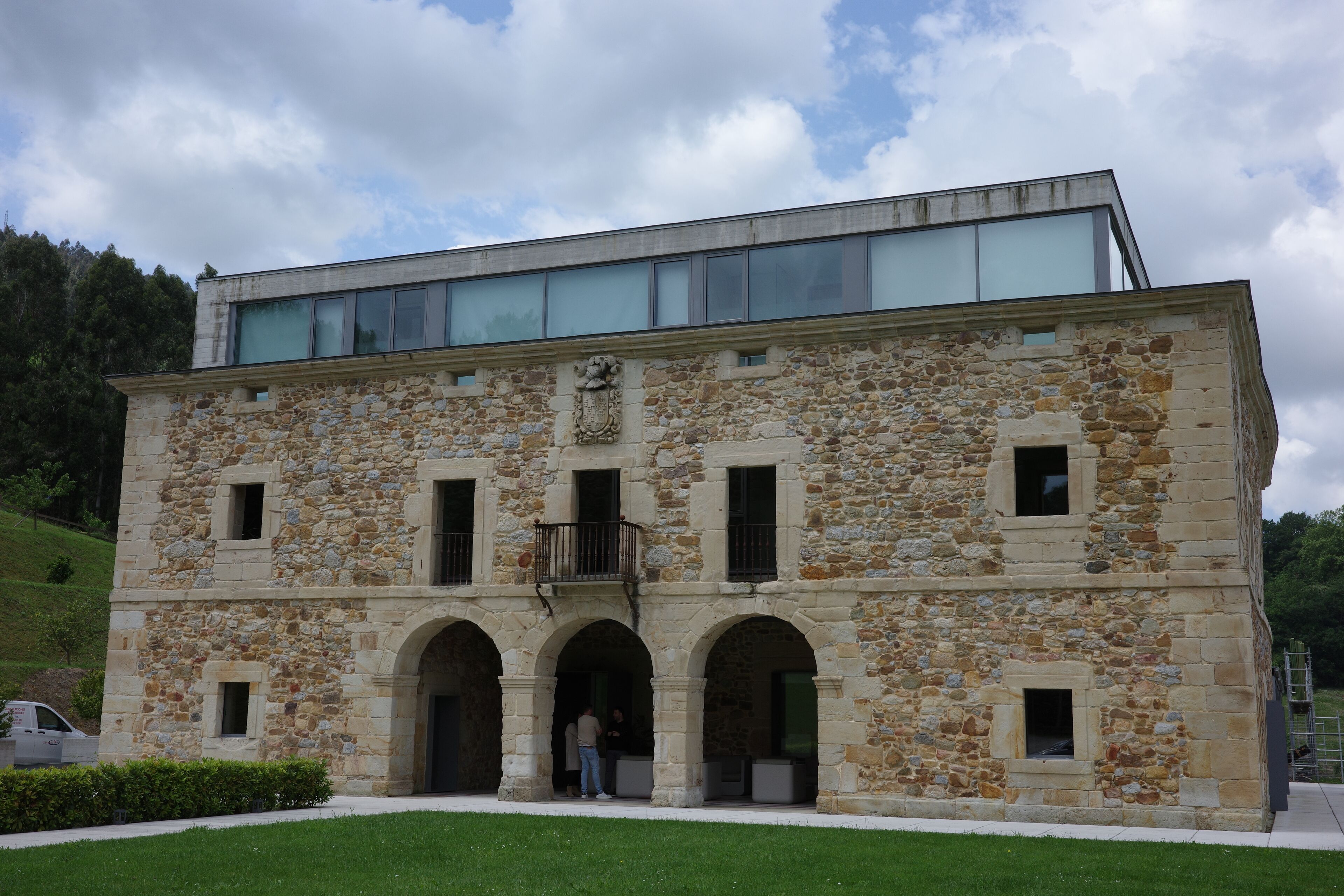 A stone building with a modern glass extension, featuring arched entrances and a balcony.
