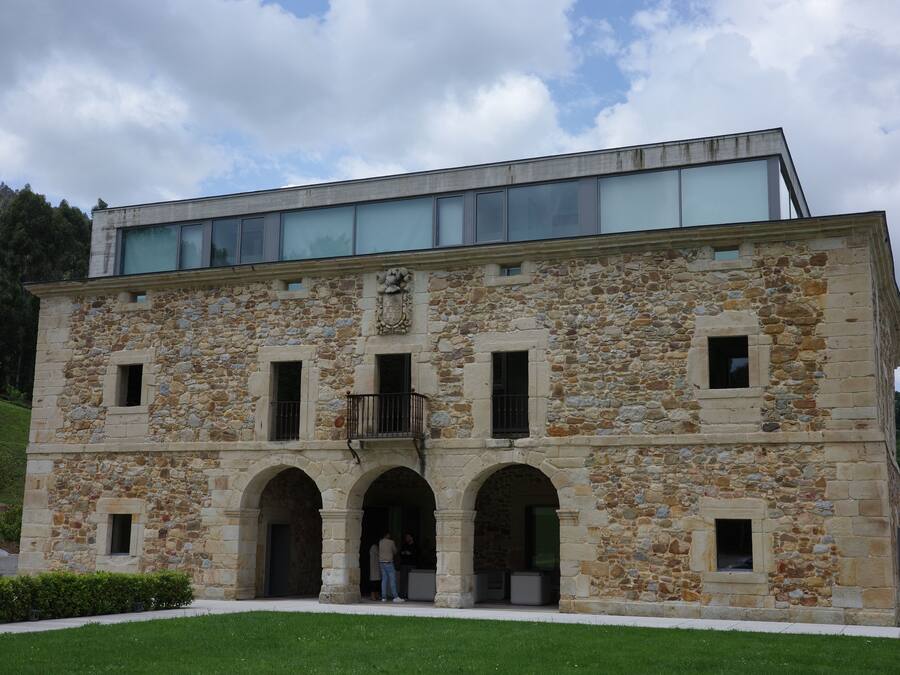 Un edificio de piedra con una extensión moderna de vidrio, que presenta entradas arqueadas y un balcón.