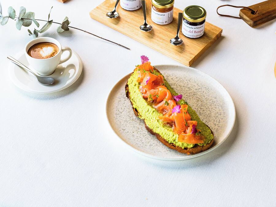 A table with a plate of avocado toast, a cup of coffee, and several condiments on wooden boards.