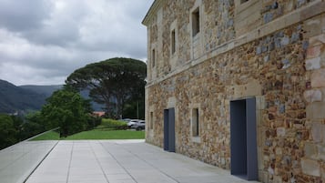 Un bâtiment en pierre avec un chemin pavé, un grand arbre et une chaîne de montagnes en arrière-plan.