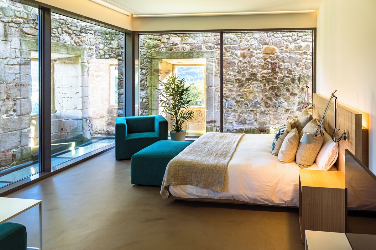 Un dormitorio moderno con pared de piedra, una cama grande, un sillón azul y vistas al exterior.