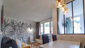 Een moderne woonkamer met een bank, een glazen salontafel, een eettafel met stoelen, een groot raam en een muur met een bladmotief.