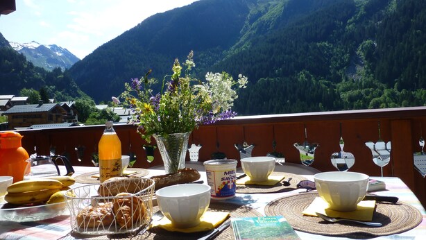 A table set with breakfast items, a bottle, and a book on a balcony with a mountain view.