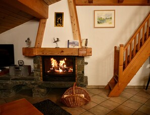 A cozy living room with a stone fireplace, wooden beams, a television, and a staircase.