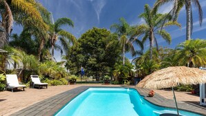Ein Pool mit Liegestühlen und einem Reetdachschirm, umgeben von Palmen und blauem Himmel.