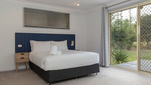 Ein Schlafzimmer mit einem großen Bett, einem Nachttisch, einer Wandleuchte und Blick auf Grünflächen durch eine Schiebetür.
