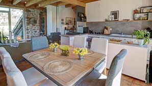 A cozy dining area with a wooden table, woven placemats, and chairs. A kitchen with modern appliances and a stone wall is visible in the background. - Charming cottage near ski trails, 2 bedrooms (Saint-Régis-du-Coin)