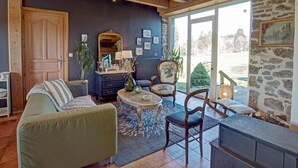 A cozy living room with a wooden ceiling, a stone wall, and a large window. There is a green sofa, a wooden chair, and a small table with a vase. - Charming cottage near ski trails, 2 bedrooms (Saint-Régis-du-Coin)