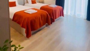 Habitación de hotel con dos camas, cada una con colcha roja y almohadas blancas, una lámpara de pie y un ventanal con cortinas traslúcidas.