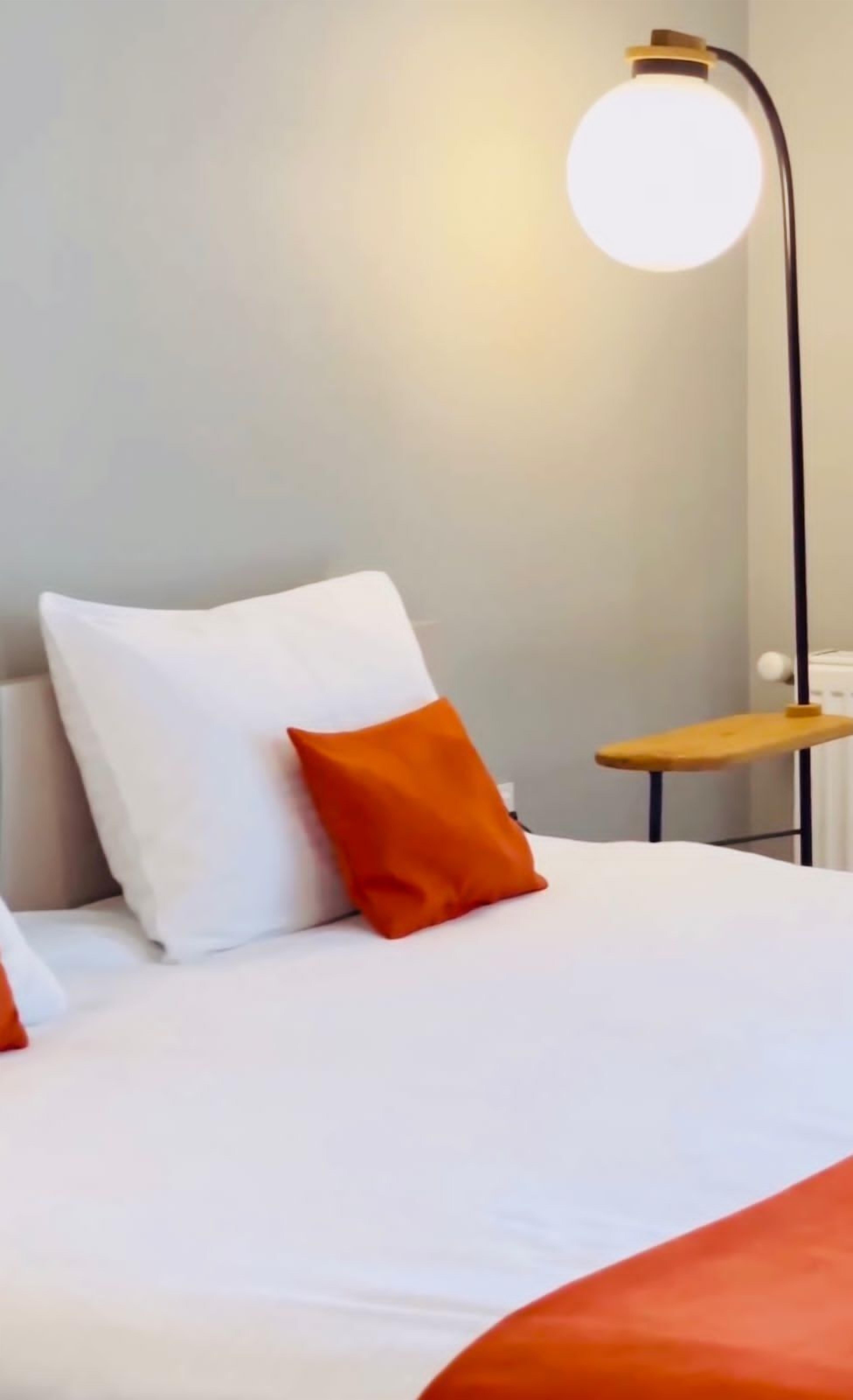 Una cama bien hecha con almohadones blancos y naranja, una mesita de noche de madera y una lámpara de pie alta con una pantalla redonda.