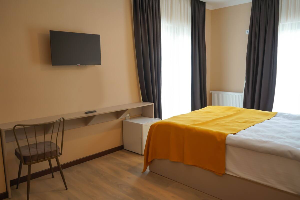 Quarto de hotel com uma cama, uma secretária com cadeira, televisão na parede e janela com cortinas.
