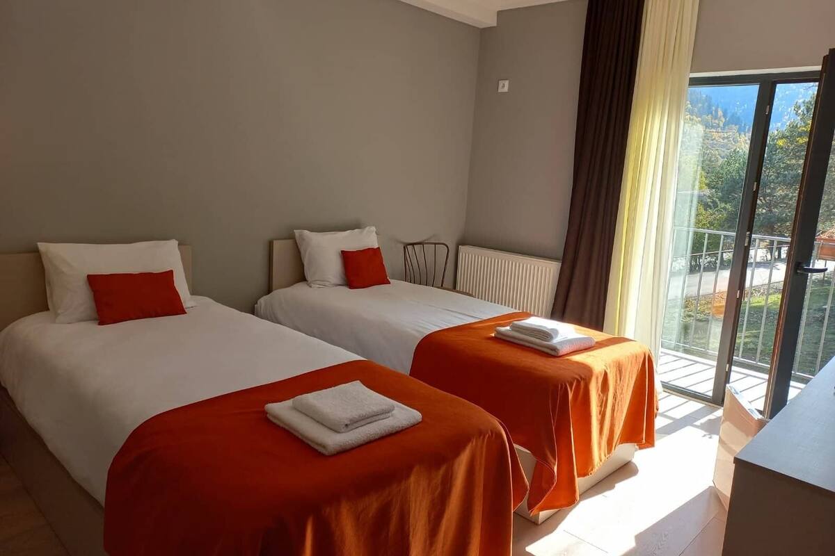 Quarto de hotel com duas camas, cada uma com roupa de cama branca e almofadas vermelhas, uma janela grande com vista para vegetação, e uma varanda com guarda-corpo.