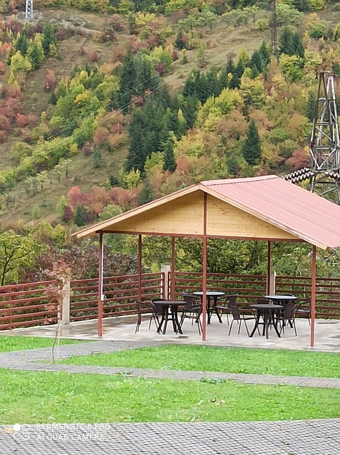 Un espacio de estar al aire libre cubierto con mesas y sillas, rodeado por una valla de metal, con un paisaje montañoso de otoño al fondo.