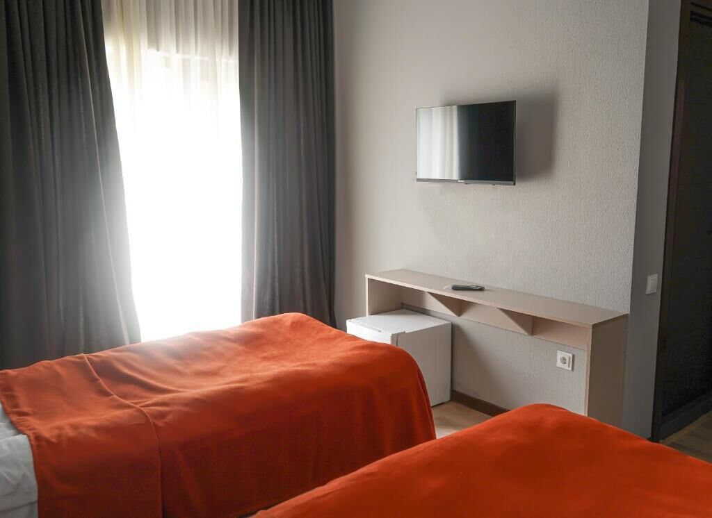 Una habitación de hotel con dos camas, un televisor montado en la pared, un escritorio pequeño y una ventana con cortinas.