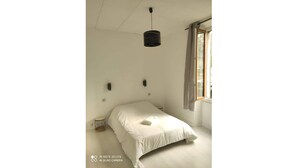 Une chambre à coucher au style minimaliste, avec un lit simple, un luminaire suspendu, une petite table de chevet et une fenêtre avec des rideaux.