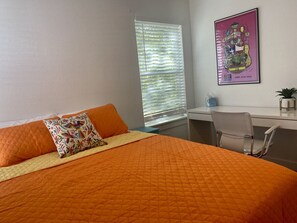 A bedroom with a large bed, an orange quilted bedspread, two pillows, a desk with a chair, a window with blinds, and a framed picture on the wall.