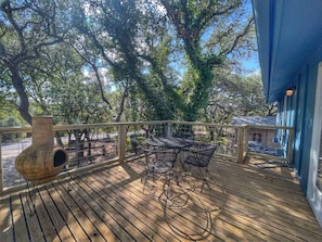 Une terrasse en bois avec une table et des chaises, un pot de fleurs suspendu, et une vue sur des arbres et un bâtiment.