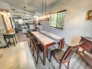 Un vaste espace dédié à la salle à manger, avec une longue table en bois et des chaises, une cuisine en arrière-plan et une fenêtre équipée de stores.
