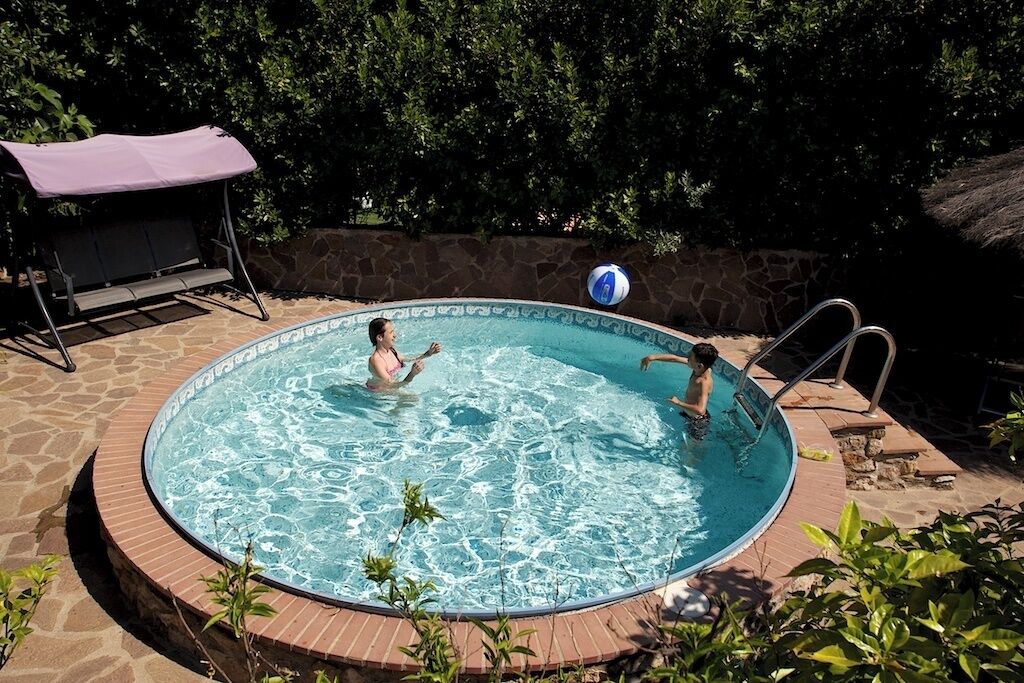 Una piscina circular con borde de piedra, rodeada de vegetación y un área de estar cubierta.