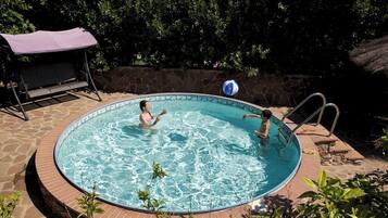 Una piscina circular con borde de piedra, rodeada de vegetación y un área de estar cubierta.