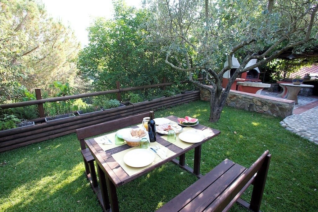 Un área de comedor al aire libre con una mesa de madera dispuesta para dos personas, rodeada por un cerco de madera y abundante vegetación.