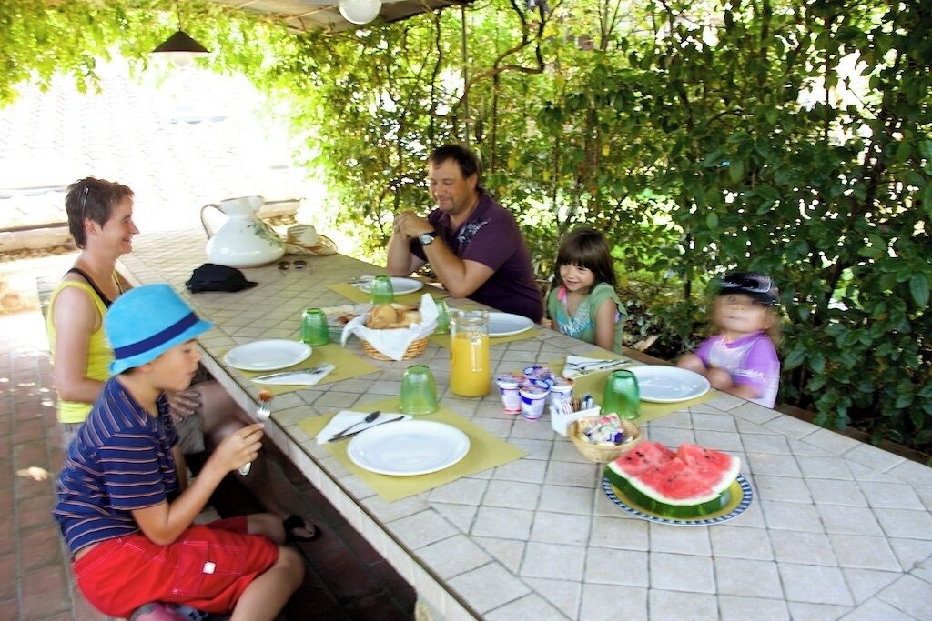 Une famille est assise à une table à l’extérieur, où elle prend son repas. On voit des assiettes, une tranche de pastèque et diverses boissons.
