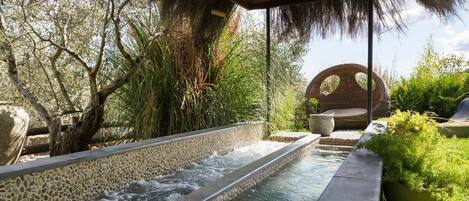 A tranquil outdoor pool area with a stone-lined water feature, surrounded by lush greenery and a wooden pergola.