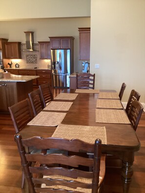 A dining area with a long wooden table and matching chairs, a kitchen with wooden cabinets and stainless steel appliances, and a tiled backsplash.