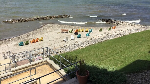 A beach with a row of colorful lounge chairs and a small wooden table, a grassy area, and a rocky shoreline with waves.