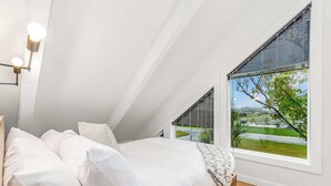 A bedroom with a large bed, white bedding, a bedside lamp, and a view of a green landscape through the window.