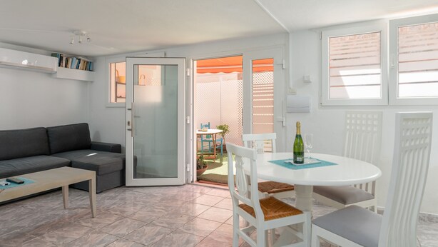 Una sala de estar con un sofá, una mesa de centro, una mesa de comedor con sillas y un ventanal corredizo que da a un espacio exterior con una mesa y sillas.