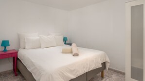 Una cama bien hecha con sábanas blancas y una toalla enrollada, una mesita de noche rosa con una lámpara azul turquesa, y una vista del exterior a través de una ventana.