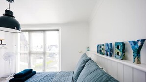 A bedroom with a bed, a wall-mounted shelf with decorative letters, a window with blinds, and a ceiling fan.