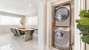 A modern laundry room with two stacked washers and dryers, a wooden table with chairs, and a potted plant. - Retreat Near MIA • Playground & Heated Pool, BBQ (Miami)