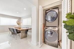 A modern laundry room with two stacked washers and dryers, a wooden table with chairs, and a potted plant. - Retreat Near MIA • Playground & Heated Pool, BBQ (Miami)