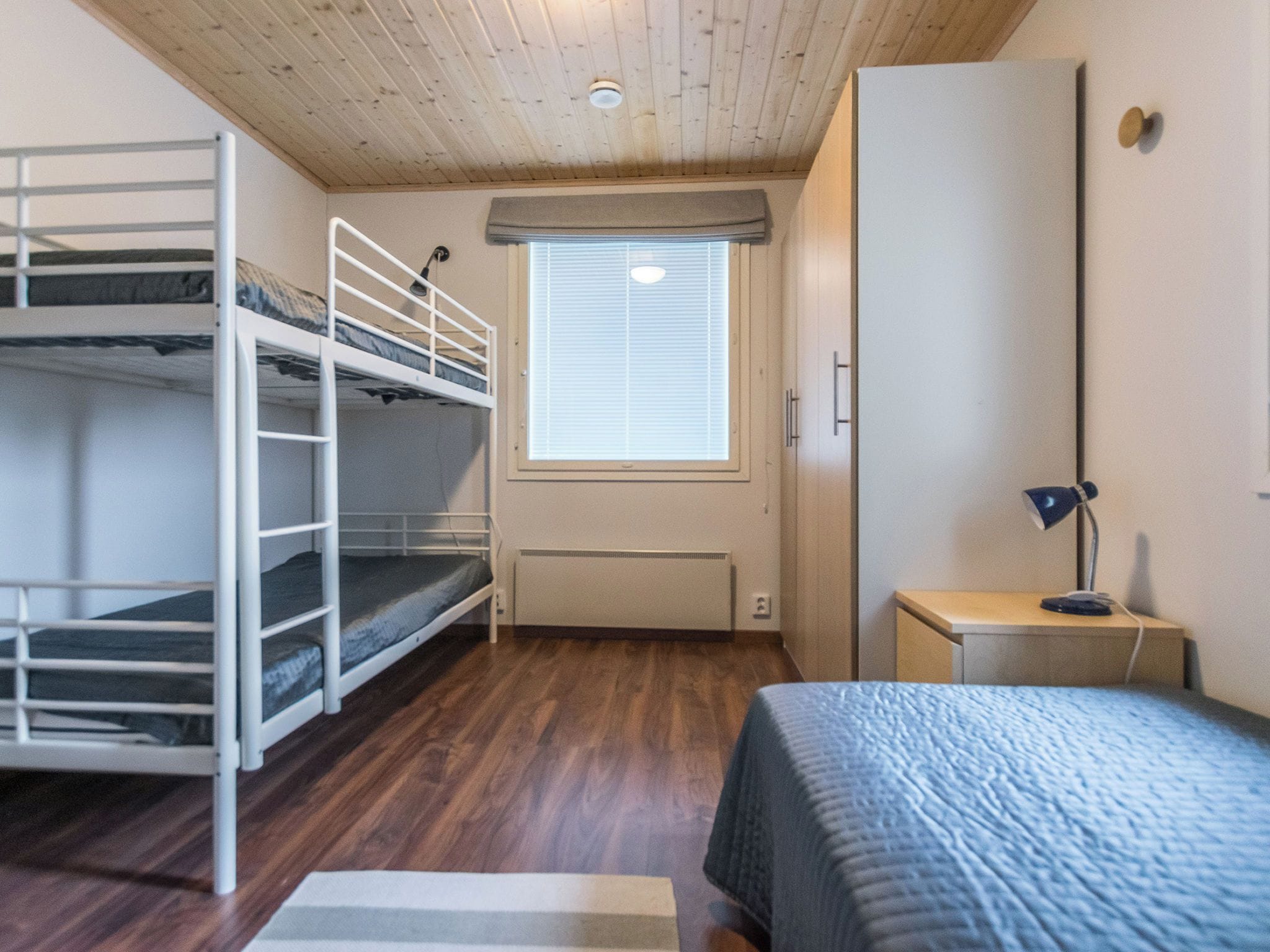 A dormitory room with bunk beds, a radiator, a window with blinds, a desk with a lamp, and a wardrobe.