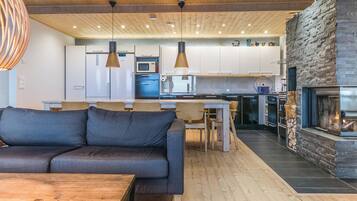 A modern kitchen with a stone fireplace, wooden ceiling, and pendant lighting.