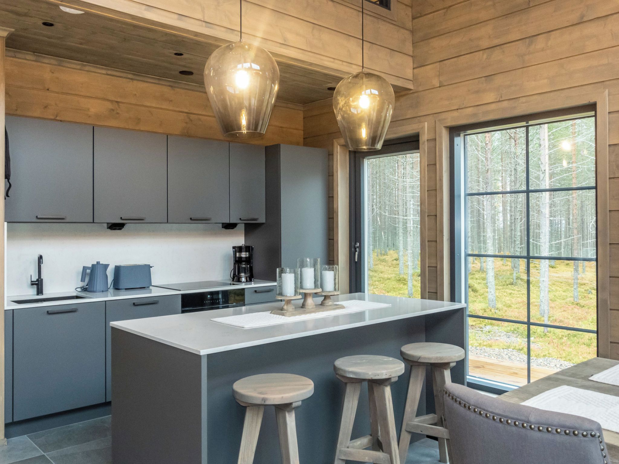 A modern kitchen with grey cabinets, a central island with stools, and a large window offering a forest view.