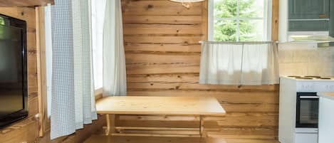 A wooden cabin interior with a wooden table and bench, a television, a window with curtains, and a kitchen area with a stove.
